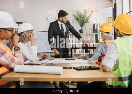 Eine Gruppe von fünf multirassischen Menschen, die formelle Kleidung und Helme tragen und ein gemeinsames Projekt im Büro erstellen und planen. Partner, die in Innenräumen mit Skizzen arbeiten. Stockfoto