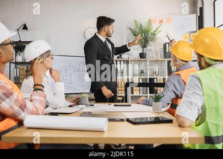 Eine Gruppe von fünf multirassischen Menschen, die formelle Kleidung und Helme tragen und ein gemeinsames Projekt im Büro erstellen und planen. Partner, die in Innenräumen mit Skizzen arbeiten. Stockfoto