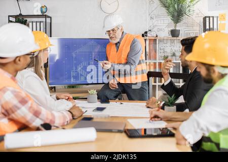 Reifer Baumeister kaukasischer Mann spricht über das Projekt auf einem großen lsd-Bildschirm mit Blaupausen an seine Kollegen Architekten, Bauherren und Designer. Teamarbeit und Brainstorming-Konzept. Stockfoto