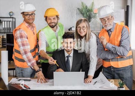 Designer, Architekten und Ingenieure treffen sich im Büro, um ein Projekt für ein neues Gebäude zu erstellen. Partner unterschiedlichen Alters, Geschlechts und Rassen stehen in der Nähe oder sitzen am Tisch und arbeiten mit Blaupausen Stockfoto