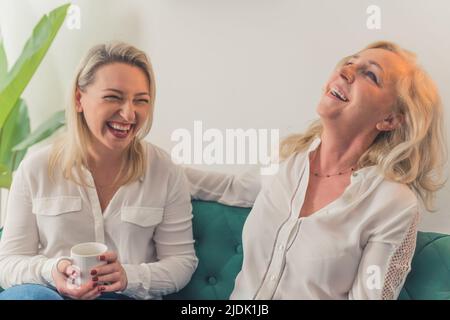 Nahaufnahme in einer Wohnung von zwei blonden, attraktiven Frauen aus den Jahrtausenden, die weiße, elegante Blusen tragen, lachen hart. Hochwertige Fotos Stockfoto