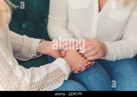 Supportkonzept. Zwei kaukasische Frauen in weißen Hemden und Denim-Hosen sitzen auf einem Sofa und halten sich die Hände. Hochwertige Fotos Stockfoto