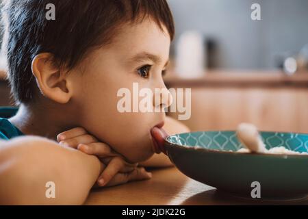 Kaukasischer Junge sitzt auf dem Küchentisch und zeigt Zunge und will nicht essen. Gesunde Ernährung, Ernährung. Familienbetreuung. Gesunder Lebensstil. Stockfoto