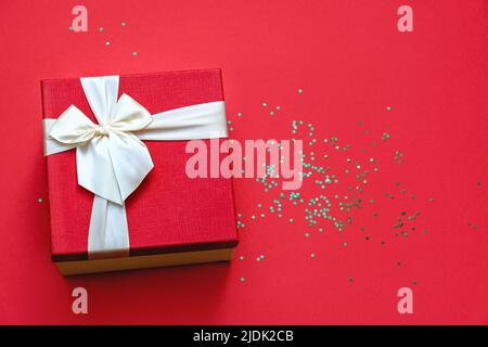 Weicher Fokus und Draufsicht auf schöner roter Geschenkbox mit Satinlichtband auf rotem Hintergrund. Grüne Glitzersterne leuchten auf dem Tisch. Geschenk zu Weihnachten und Neujahr. Platz für Text kopieren. Stockfoto