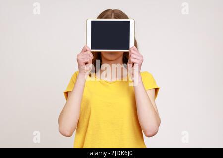 Porträt einer unbekannten Frau in gelbem Casual-Stil T-Shirt-Abdeckung Gesicht mit Digital-Tablet mit leerem Bildschirm, Kopierer Platz für Werbung. Innenaufnahme des Studios isoliert auf grauem Hintergrund. Stockfoto