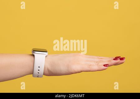 Nahaufnahme der Hand der Frau mit roter Maniküre und Armbanduhr mit weißem Armband, Technologie-Gerät. Innenaufnahme des Studios isoliert auf gelbem Hintergrund. Stockfoto