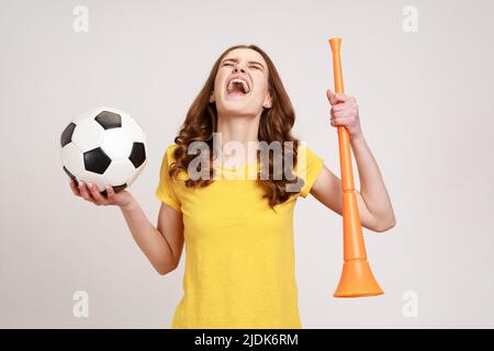 Aufgeregt Teenager-Mädchen in gelbem T-Shirt mit Horn und schwarz-weißen Fußball, den Sieg der Lieblings-Fußballmannschaft in der Meisterschaft zu feiern. Innenaufnahme des Studios isoliert auf grauem Hintergrund. Stockfoto