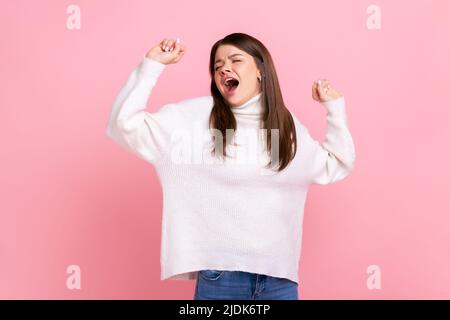 Verschlammte junge Frau, die mit geschlossenen Augen gähnend steht, die Arme streckt, sich erschöpft fühlt und sich ausruhen muss, trägt einen weißen Pullover im lässigen Stil. Innenaufnahme des Studios isoliert auf rosa Hintergrund. Stockfoto