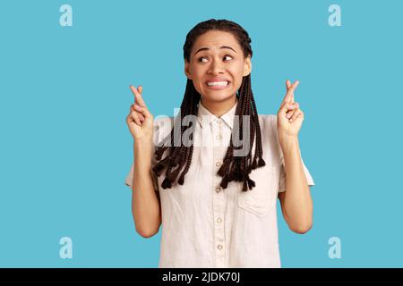 Junge Frau mit schwarzen Dreadlocks macht Wunsch glaubt an Glück, hofft, dass Träume wahr werden, hat hoffnungsvollen Ausdruck, erwartet etwas. Innenaufnahme des Studios isoliert auf blauem Hintergrund. Stockfoto