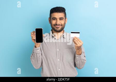 Fröhlicher bärtiger Geschäftsmann mit leerem Display, Smartphone und Kreditkarte, zufrieden mit einfachem Mobile Banking, im gestreiften Hemd. Innenaufnahme des Studios isoliert auf blauem Hintergrund. Stockfoto