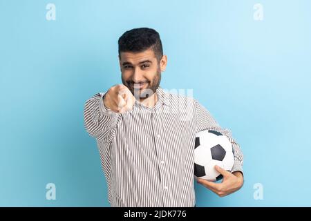 Porträt eines jungen erwachsenen bärtigen Geschäftsmannes, der auf Sie zeigt, den Fußball hält, die Kamera mit fröhlicher Optik anschaut und ein gestreiftes Hemd trägt. Innenaufnahme des Studios isoliert auf blauem Hintergrund. Stockfoto