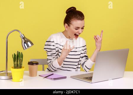 Aufgeregt freche Bloggerin zeigt Rock and Roll Finger Geste Blick auf Laptop-Bildschirm, Chat Web-Kamera sitzen am Arbeitsplatz. Studio-Innenaufnahme isoliert auf gelbem Hintergrund. Stockfoto