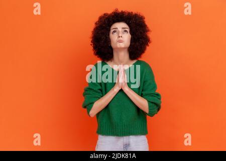 Hoffnungsvolle Frau mit Afro-Frisur in grünem Pullover, die die Hände im Gebet hält und fleht, mit hoffnungsvollen Augen aufschaut und sich entschuldigt. Innenaufnahme des Studios isoliert auf orangefarbenem Hintergrund. Stockfoto