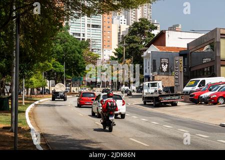 Autos, die an einem gekrümmten Abschnitt der Sumare Avenue in der Nähe einiger kleiner Geschäfte im Viertel Perdizes an einem normalen Arbeitstag unter sonnenklarem, blauem Himmel vorbeifahren. Stockfoto