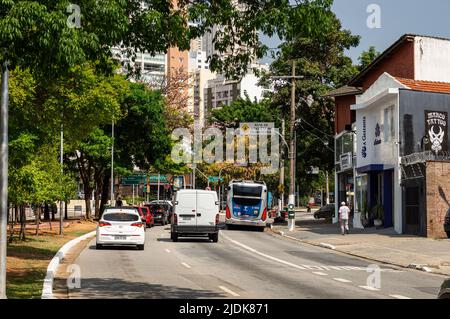 Autos, die an einem gekrümmten Abschnitt der Sumare Avenue im Viertel Perdizes vorbeifahren, in der Nähe von kleinen lokalen Geschäften an einem normalen Arbeitstag unter sonnigem, blauem Himmel. Stockfoto