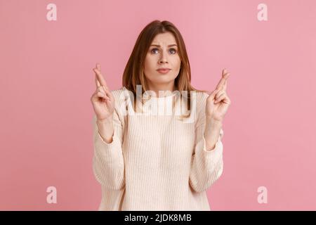 Portrait einer blonden Frau, die gekreuzte Daumen hält und sich viel Glück wünscht, aufgeregt aussieht, auf den Sieg hofft, weißen Pullover trägt. Innenaufnahme des Studios isoliert auf rosa Hintergrund. Stockfoto