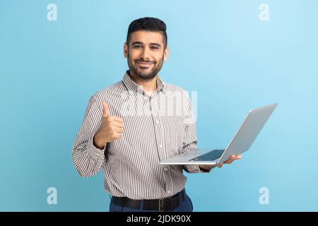 Porträt eines lächelnden, positiven Geschäftsmannes, der mit einem tragbaren Computer in der Hand steht, auf das Display schaut, den Daumen nach oben zeigt und ein gestreiftes Hemd trägt. Innenaufnahme des Studios isoliert auf blauem Hintergrund. Stockfoto