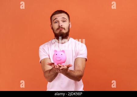 Porträt eines traurig frustrierten bärtigen Mannes, der Sparschwein in den Händen hält, unglücklich ist, keine Ersparnisse hat, Trauer ausdrückt und ein rosa T-Shirt trägt. Innenaufnahme des Studios isoliert auf orangefarbenem Hintergrund. Stockfoto