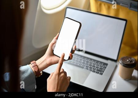 Nahaufnahme, weibliche Reisende mit einem Smartphone und einem tragbaren Laptop im Flugzeug. Modell mit weißem Bildschirm für Telefon und Laptop. Stockfoto