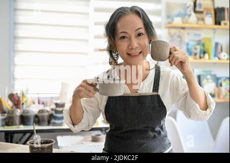 Schöne kreative asiatische gealterte Frau Töpfer zeigt ihre Keramik-Kaffeetasse, stehen in ihrem Handwerk Studio Arbeitsplatz. Stockfoto