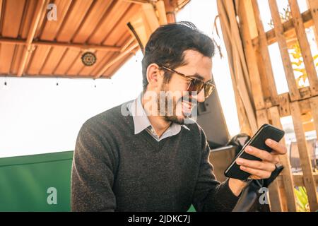 Junger bärtiger Hipster lächelt und schaut in einer Bar auf sein Handy Stockfoto