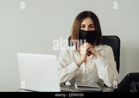 Porträt einer jungen weiblichen Büroleiterin, die in die Kamera schaut, selbstbewusste Frau in Schutzmaske, die am Arbeitsplatz sitzt Stockfoto