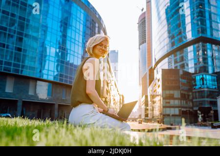 Porträt einer Frau mittleren Alters in grüner, weißer Hose, die im Stadtzentrum in der Nähe von Wolkenkratzern sitzt und einen Laptop hält. Stockfoto