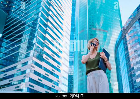 Porträt einer geschäftigen Geschäftsfrau, die im Stadtzentrum in der Nähe von Wolkenkratzern steht, einen Laptop hält und auf dem Smartphone spricht. Stockfoto