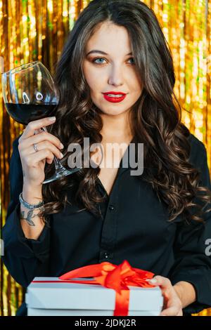 Porträt einer jungen Frau, die ein Glas Rotwein hebt, mit einer weißen Geschenkbox mit rotem Band auf einem goldenen Lametta-Hintergrund. Stockfoto