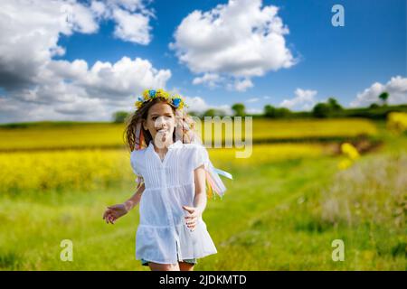 Schönes, energiegeladenes, verträumtes Teenager-Mädchen in einem weißen Kleid und einem leuchtend ukrainischen Kranz, das durch gelb blühende Rapsfelder und grüne Äcker läuft Stockfoto