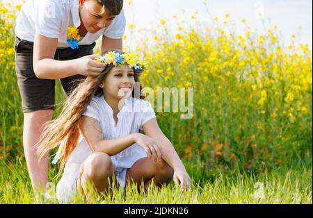 Der Bruder des Teenagers flechzt mehrfarbige Bänder in einem gelb-blauen Kranz mit wilden Blumen auf dem Kopf der fröhlich lächelnden Schwester auf der grünen Wiese A Stockfoto