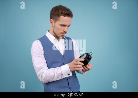 Der junge Geschäftsmann steht mit einem Wecker in den Händen auf einem blauen Studiohintergrund. Das Konzept der richtigen Zeit Stockfoto