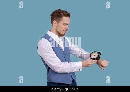 Der junge Geschäftsmann steht mit einem Wecker in den Händen auf einem blauen Studiohintergrund. Das Konzept der richtigen Zeit Stockfoto