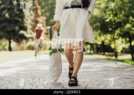 Unverkennbare Frau hält Öko-Einkaufstasche im Freien, während sie am sonnigen Sommertag im Park spazieren geht Stockfoto