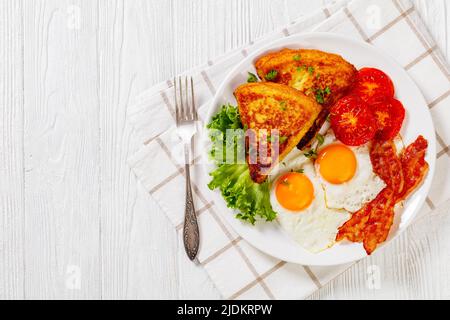 irisches Frühstück mit Kartoffelpüree, Spiegeleiern, Speckscheiben, gegrillten Tomaten und frischem Salat auf weißem Teller auf weißem Holztisch mit Besteck Stockfoto