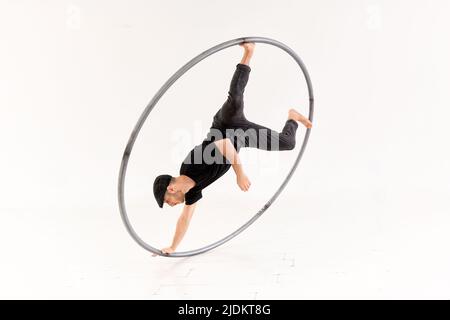 Ganzer Körper des aktiven Barfuß-akrobaten, der einen Arm Münzstunt mit cyr-Rad vor weißem Hintergrund im hellen Studio durchführt Stockfoto