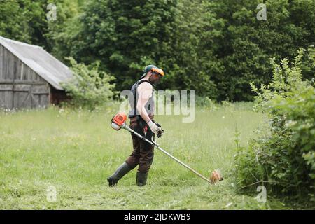 Mann mäht hohes Gras mit Benzin Rasentrimmer im Garten oder Hinterhof. Prozess des Rasentrimmens mit dem Handmäher Stockfoto