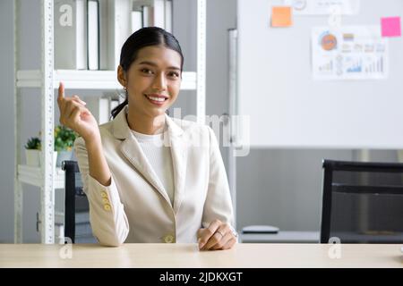 Junge asiatische Geschäftsfrau in beigefarbenen Anzug sitzen entspannen mit Smiley Gesicht im Büro. Auf dem Whiteboard befand sich ein Notizbuch und ein Dokument im Hintergrund Stockfoto