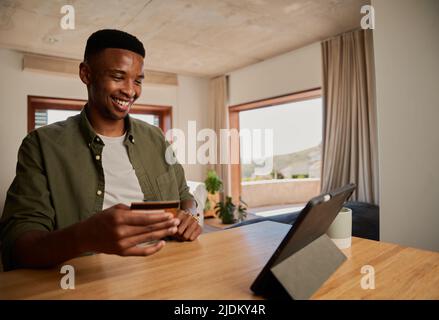 Glücklicher junger erwachsener afroamerikanischer schwarzer Mann mit elektronischem Tablet, begeistert, mit seiner Kreditkarte zu Hause am Online-Kauf zu tätigen Stockfoto