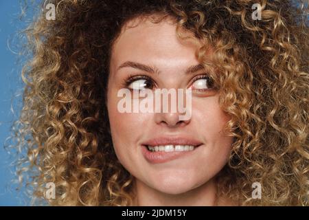 Junge blonde Frau mit lockigen Haaren lächelt und schaut isoliert auf blauen Hintergrund zur Seite Stockfoto