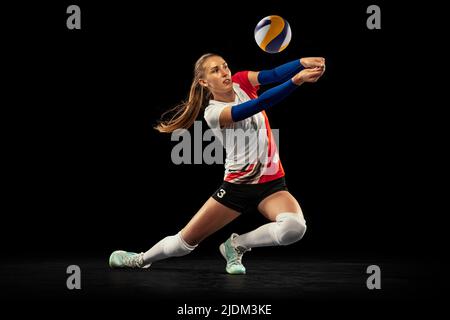 Dynamisches Porträt eines professionellen Volleyballspielers in Sportuniform, der Volleyball auf dunklem Hintergrund spielt. Sport, gesunder Lebensstil, Team Stockfoto