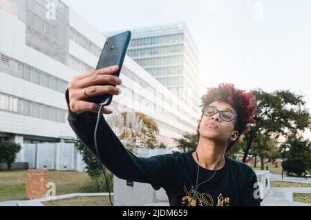 Frontansicht junge Brünette lateinkolumbianische Frau mit Afro, Brille und schwarzer Sportkleidung, im Freien ein Selfie mit ihrem Handy, auf einem öffentlichen Platz Stockfoto