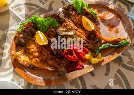 Draufsicht auf einen köstlichen, schönen gegrillten Fisch mit Pfeffer, Zitrone und grünen Salatblättern auf einem Holztablett Stockfoto