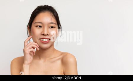 Schöne junge asiatische Frau mit glatter Haut, berührende Wange, wegschauen und lächeln, weißer Hintergrund, Freiraum Stockfoto