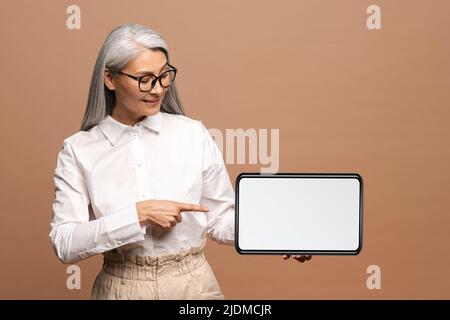 Porträt der selbstbewussten älteren Frau, die das Tablet hält und lächelnd auf den Bildschirm zeigt. Innenaufnahme des Studios auf beigefarbenem Hintergrund Stockfoto