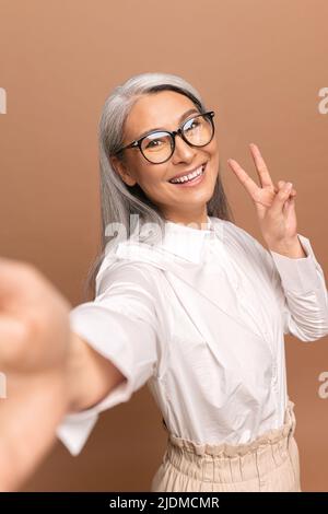 Fröhliche ältere asiatische Frau, die Selfie macht, schöne reife Frau, die die Hand in der Siegesgeste hält, lächelt und die Kamera isoliert auf Beige anschaut Stockfoto