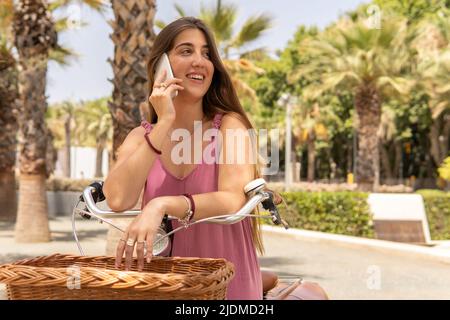 Eine junge Frau in einem hübschen rosa Kleid sitzt auf ihrem Vintage-Fahrrad und telefoniert in einem Park. Stockfoto