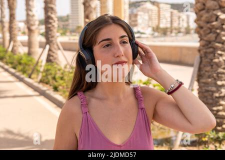 Die junge Frau in einem rosa Kleid hält und hört an einem sonnigen Tag in der Stadt Musik auf ihren schwarzen Kopfhörern. Stockfoto