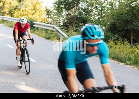 Verschwommene Bewegung von zwei Fahrern auf professionellen Rennrädern, die auf der kurvenreichen, gepflasterten Straße durch den Wald schnell bergab fahren Stockfoto