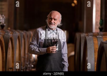 Kaukasischer Senior-Graubart-Winzer, der Rotwein verkostet, um seine Qualität und seinen Geschmack zu ermitteln, steht in der Nähe eines großen Eichenfasses Stockfoto